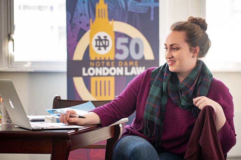 Kimberly Schreiber sits at a desk working on a laptop. A banner for the 50th anniversary of the London Law Program is behind her.