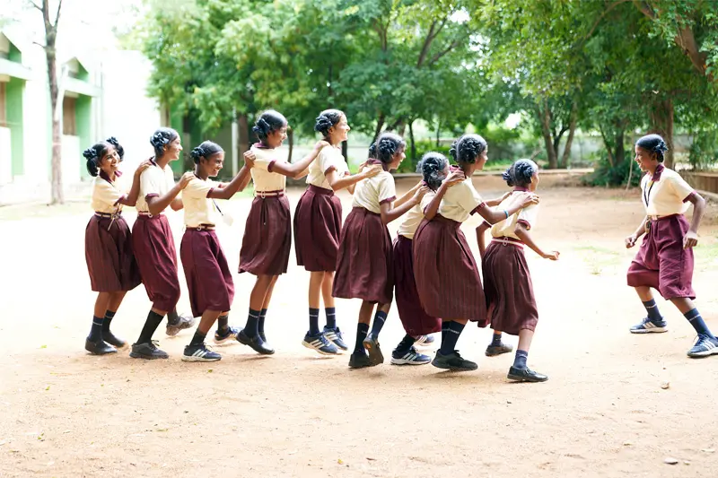 A line of Indian girls in matching tan and maroon school uniforms hold each others shoulders and playfully skip across a dirt schoolyard. Trees and a school building can be seen in the background.