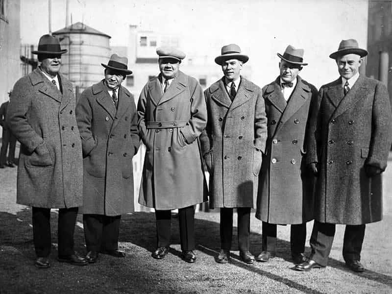 Six men pose wearing old timey hats and long peacoats.