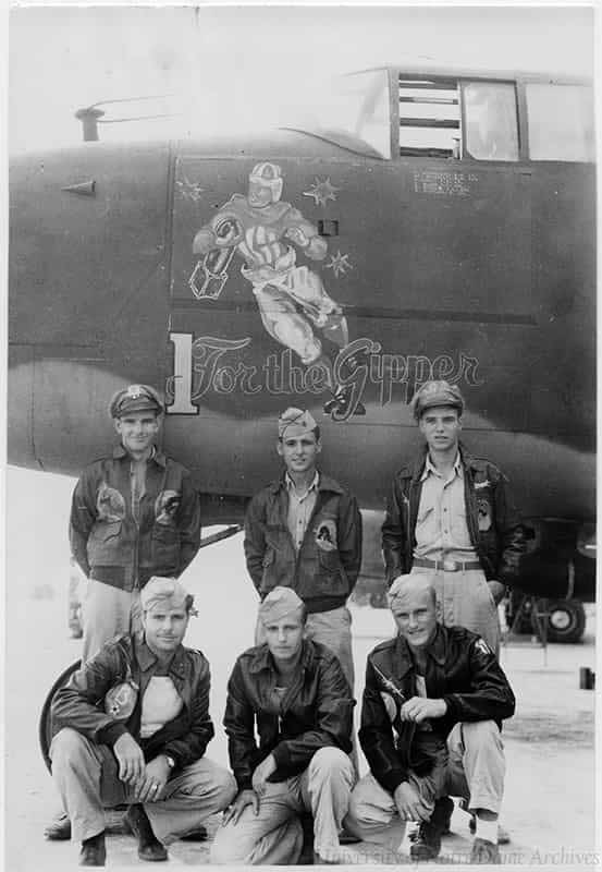Military crew in front of a World War II Bomber that has a painting of a football player holding onto a bomb that says '1 [One] for the Gipper'.