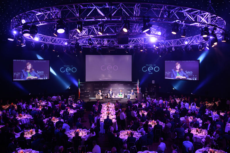 Four people sit on a stage surrounded by round tables. There are viewing screens above the stage.