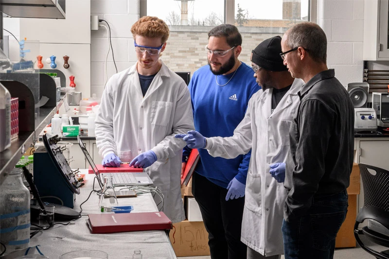 Four individuals, three wearing lab coats and gloves, gather around a lab bench, examining equipment and a small red object. One person gestures towards the equipment.