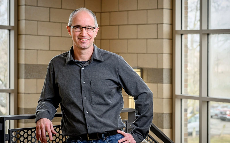 A man with short graying hair and glasses, wearing a dark gray button-down shirt and jeans, smiles as he leans against a railing by a window.