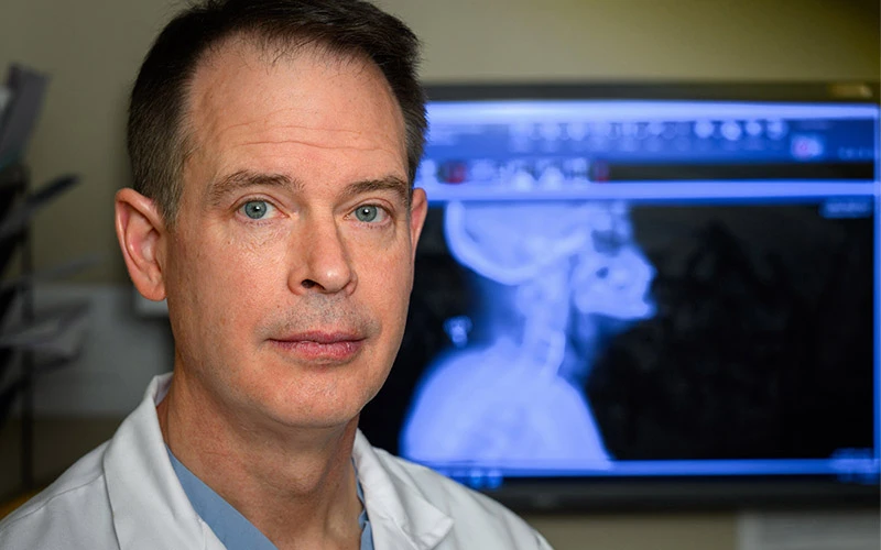 Doctor in a white coat and blue scrubs looks at the camera with a head and neck x-ray displayed on a monitor behind him.