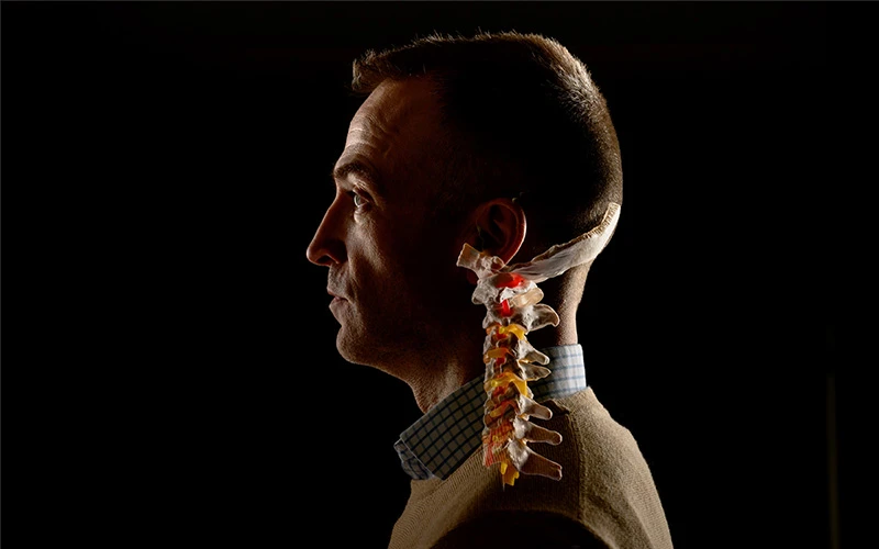 Profile of a man with short brown hair wearing a tan sweater and blue and white checked collared shirt. A model of a cervical spine section with yellow and red highlights is positioned behind his neck against a black background.