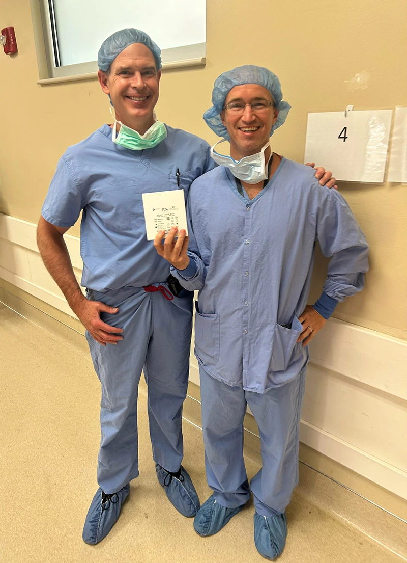 Two smiling medical personnel in blue scrubs, hair nets, and face masks stand together. One holds a small white packet. They are wearing blue shoe covers and stand next to a wall marked with the number 4.