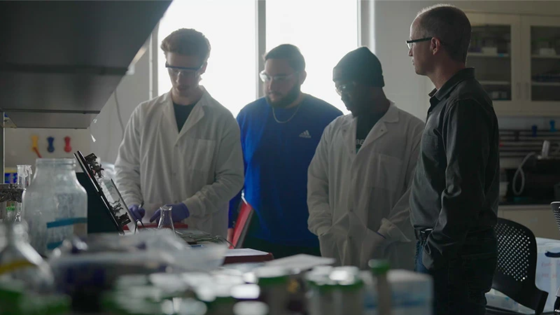 Four people in a lab, three wearing white lab coats and safety glasses, observe an experiment.  One person adjusts equipment on a red lab table containing glass beakers and other supplies. Another person wears a royal blue Adidas sweatshirt and black knit cap. A man in a dark gray, long-sleeved shirt observes with his hands in his pockets.