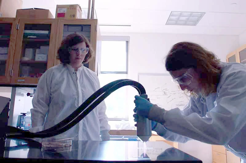 Two researchers in white lab coats and safety glasses work with scientific equipment in a lab. One researcher observes as the other directs a handheld instrument attached to a large flexible tube toward a sample on a dark table.