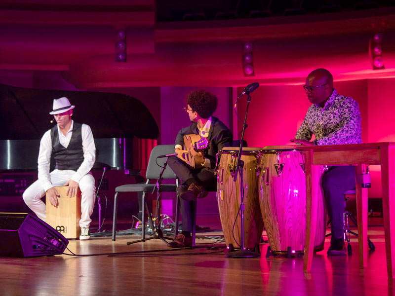 Jaime El Estampio sits on a wooden drum while Antonio Herrera plays guitar and an artist beats on hand drums.