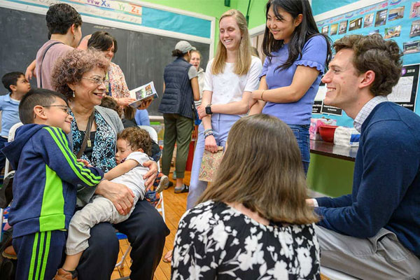 Students talk and smile with families in a classroom. An elderly woman is holding a child and another one leans on her.