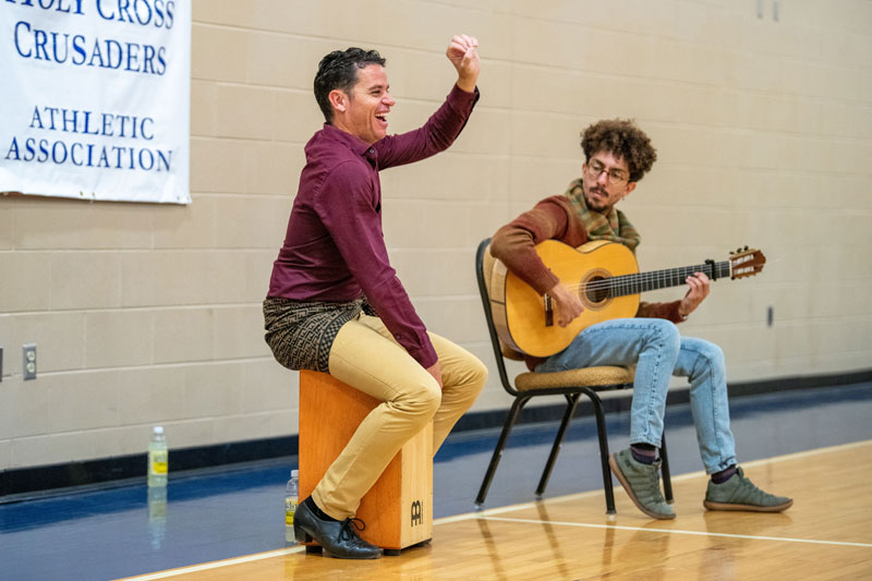 Jaime El Estampio and Antonio Hererra sit in a school gym performing on drums and acoustic guitar.