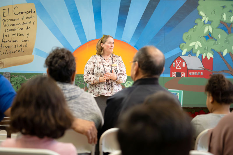 Notre Dame professor Tatiana Botero stands in front of a mural of a sunset and barn as she speaks to a group of adults and children.