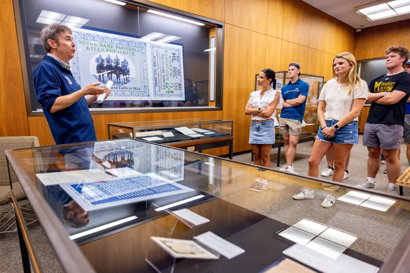 A male gestures toward a framed 1924 poster reading 'Notre Dame Football Kills Prejudice' in the University Archives exhibit. Several students observe artifacts in display cases.