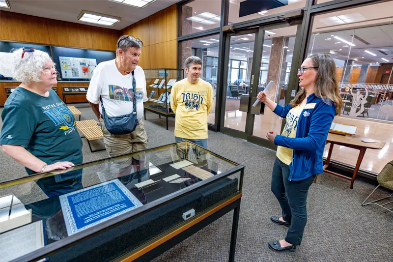 A female gestures towards glass doors while giving a tour of the library’s exhibit to three visitors. The visitors wear Notre Dame apparel and look at items in a glass display case containing historical documents.