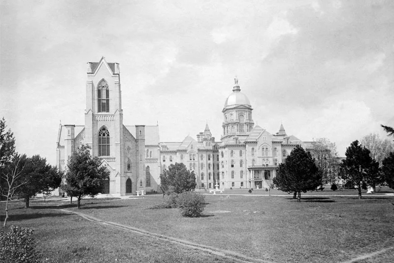 A black and white photo of the University of Notre Dame campus. The Main Building, with its iconic golden dome, stands prominently to the right. To the left, a tall, gothic-style building with a pointed spire adds architectural contrast. Trees dot the grassy area separating the buildings. Dirt paths crisscross the lawn in the foreground.