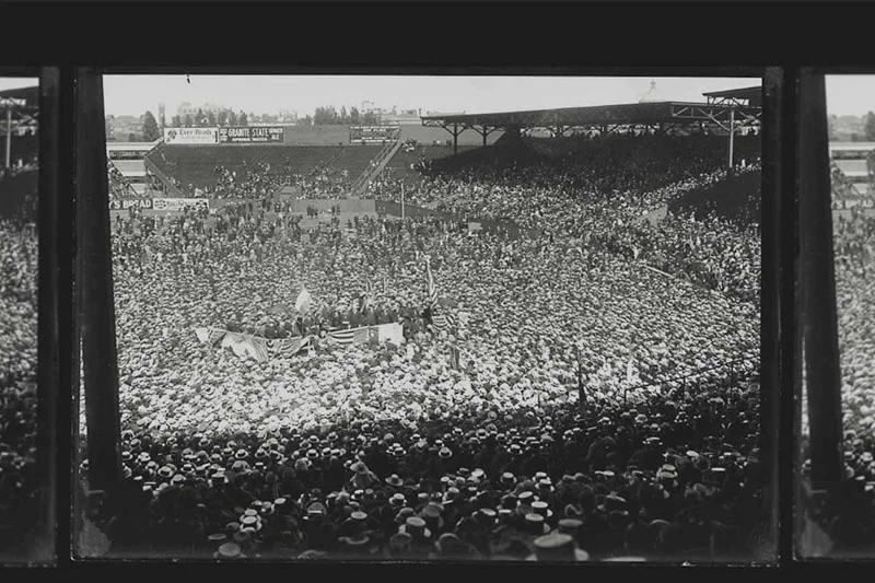Black and white photo of a massive crowd gathered in a stadium. American flags are visible in the center of the field, surrounded by a small group of people. The stands are full, and the crowd extends onto the field itself. The image is framed by the dark vertical bars from a window