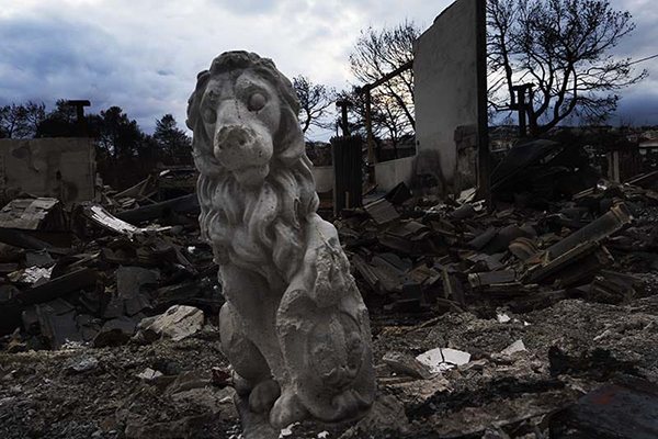 A statue of a lion is cracked and broken. It stands among the ruins of a building behind it.