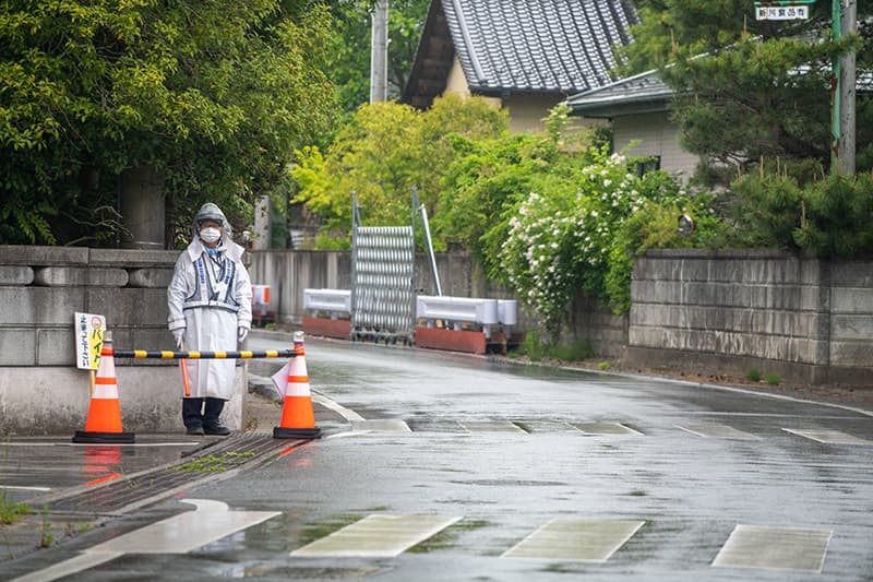 A guard in a hazmat suit and mask stands behind two orange cones alongside an evacuation zone in Futaba.