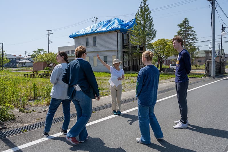 A woman wearing a hat stands in front of a two story home with a blue tarped roof. She points toward the home while four people from Notre Dame look onward.