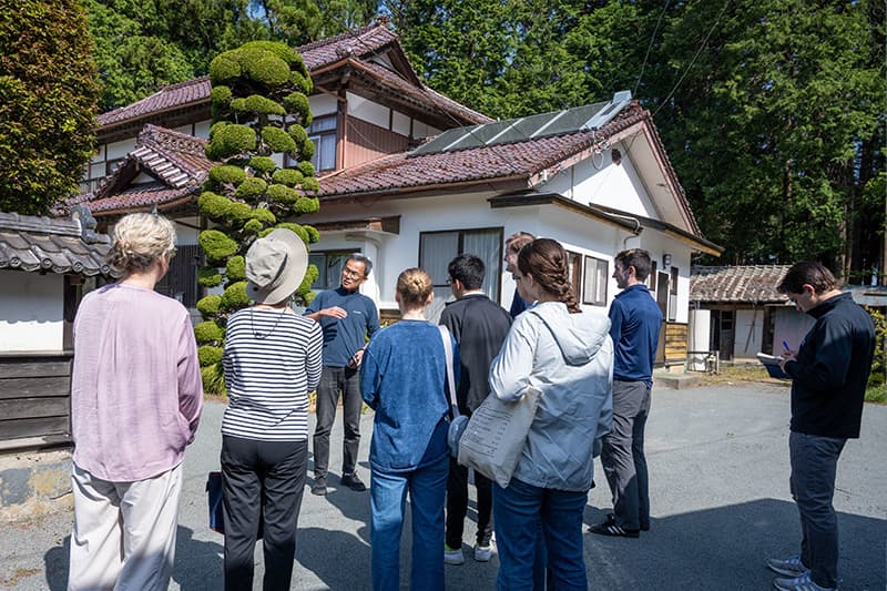 Students and staff gather around a man in front of a white two-story home with a brown terracotta roof.