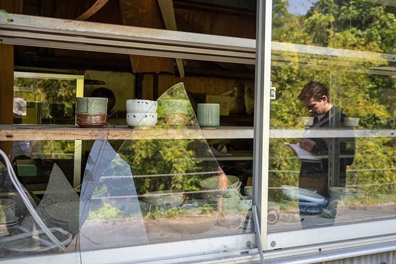 A shattered window gives way to a shelf lined with pottery. In the reflection you can see a young male with his head bent writing on a notepad.