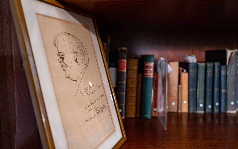 A caricature of GK Chesterton in a frame, sitting to next to a shelf with all his books.