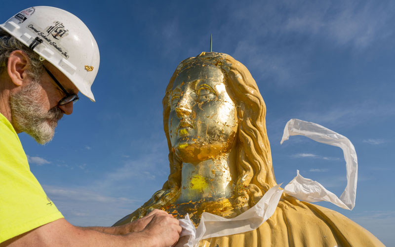 An image of a man in a hard hat applying gold to the statue of Mary on top of the Golden Dome.