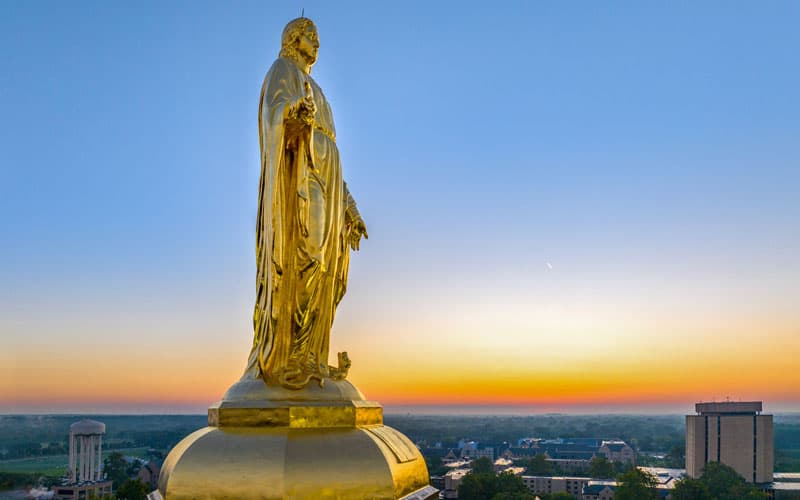A close up of the statue of Mary on top of the Golden Dome with a beautiful orange sunset in the background.