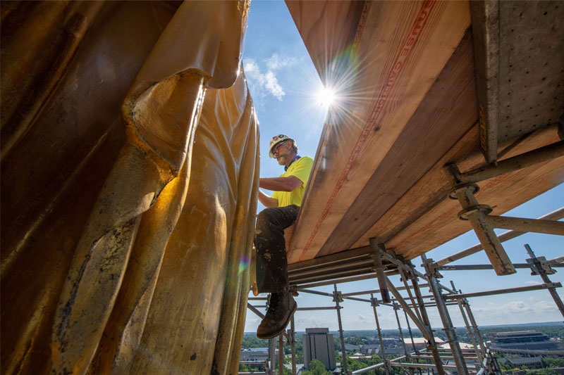 A view from below of a man sitting on scaffolding, applying gold leaf to the Golden Dome.