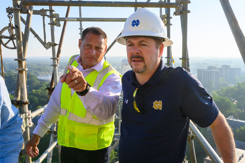 Tony Polotto stands on scaffolding pointing to something in the distance and Mike Daly looks on.