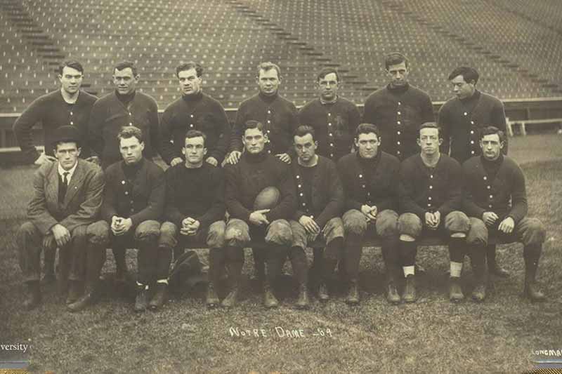 A sepia picture of 15 men on a football field in an empty stadium.