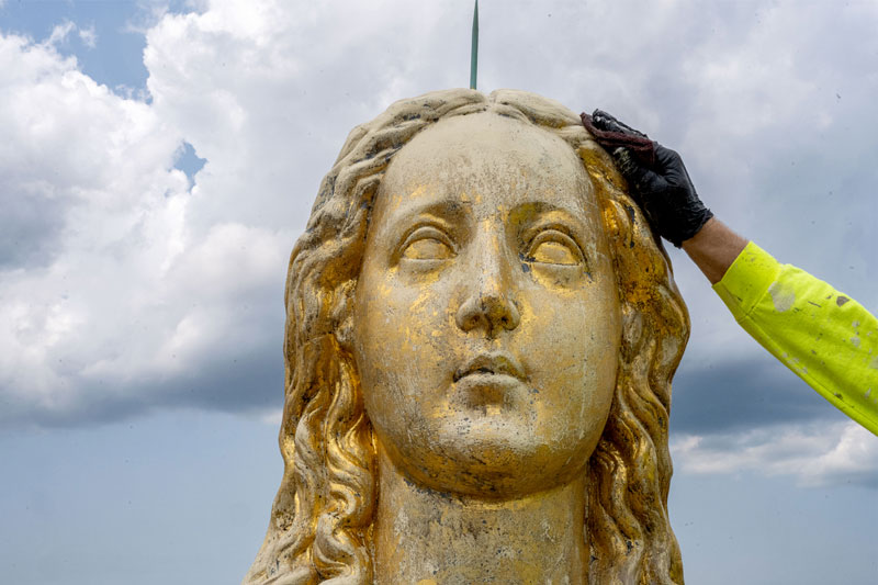 A close-up of face on the statue of Mary prior to regilding. Her face is dull instead of shiny, and you can see black coming through from underneath. A gloved hand is rubbing the surface with a soft cloth. 