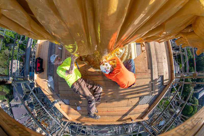 A man and a women lay on top of scaffolding at the base of the state of Mary on top of the Golden Dome. They are applying layers of gold leaf to the base.