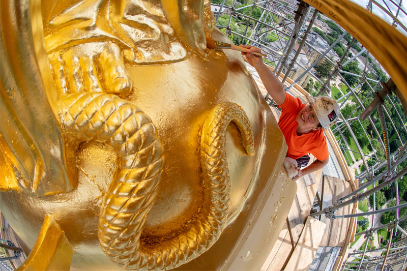 A close of view of the newly gilded snake at the base of the statue of Mary on top of the Golden Dome. In the background is a gilder holding a paint brush making final touches on the gold leaf.
