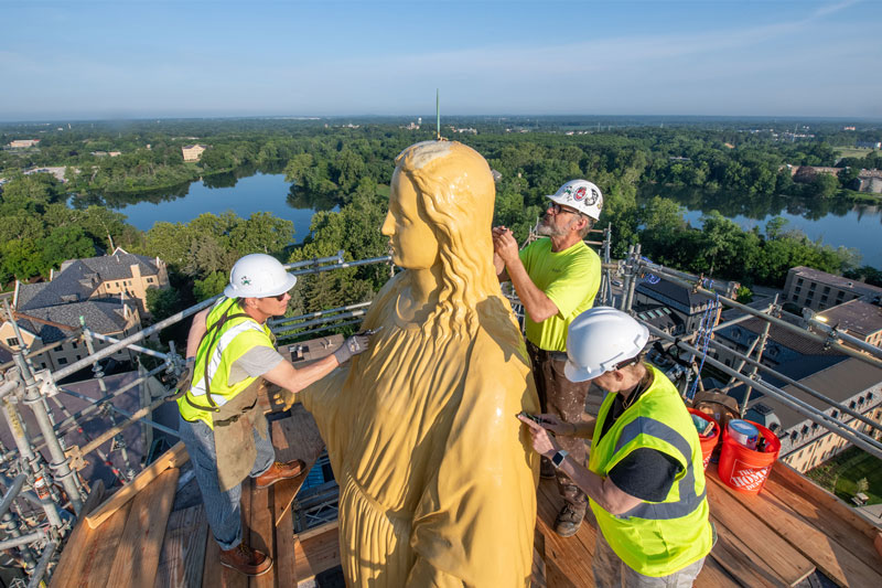 Three gilders stand on scaffolding near the statue of Mary on top of the Golden Dome. They're all working on her surface.