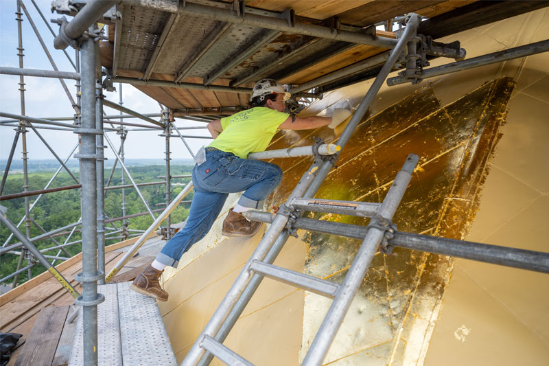 A woman stands on scaffolding as she leans on the Golden Dome, cleaning the surface with a soft cloth.