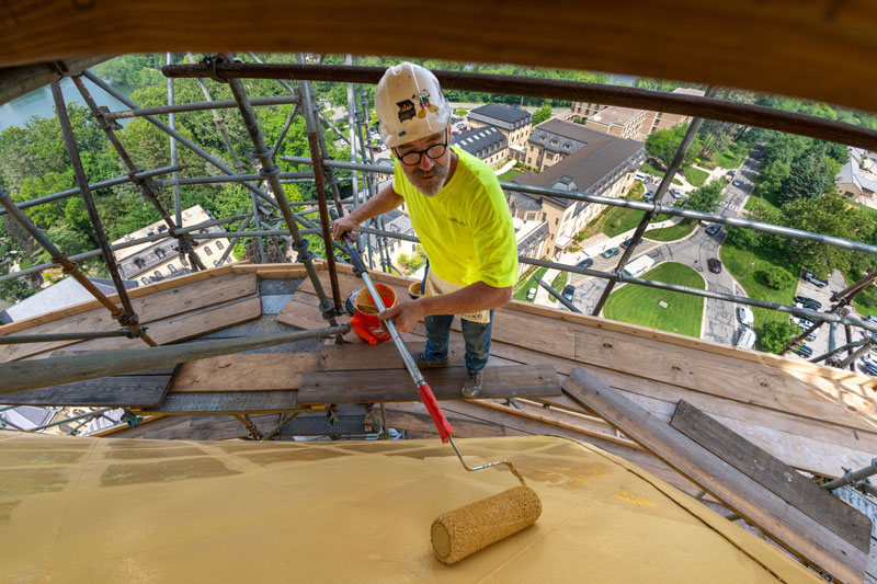 A view of a man from above. He's standing on scaffolding and rolling paint primer onto the surface of the Golden Dome. 