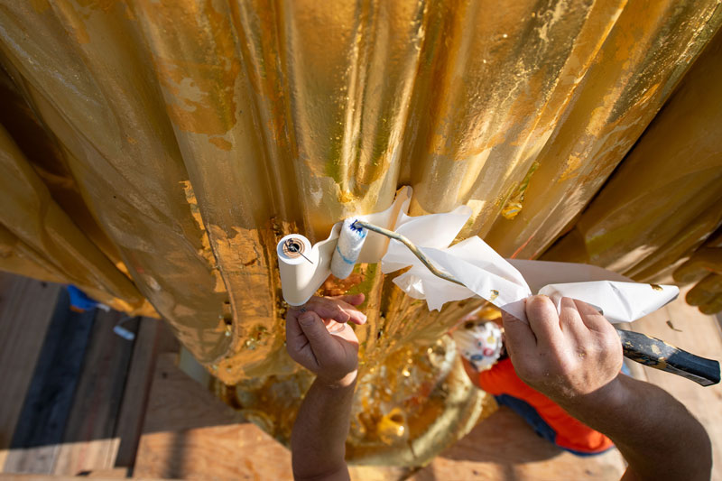 A close up view of a roll of gold leaf being applied to the base of the statue of Mary on top of the Golden Dome, by a roller.