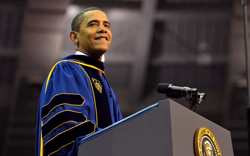 Barack Obama smiles from a podium, dressed in graduation regalia.