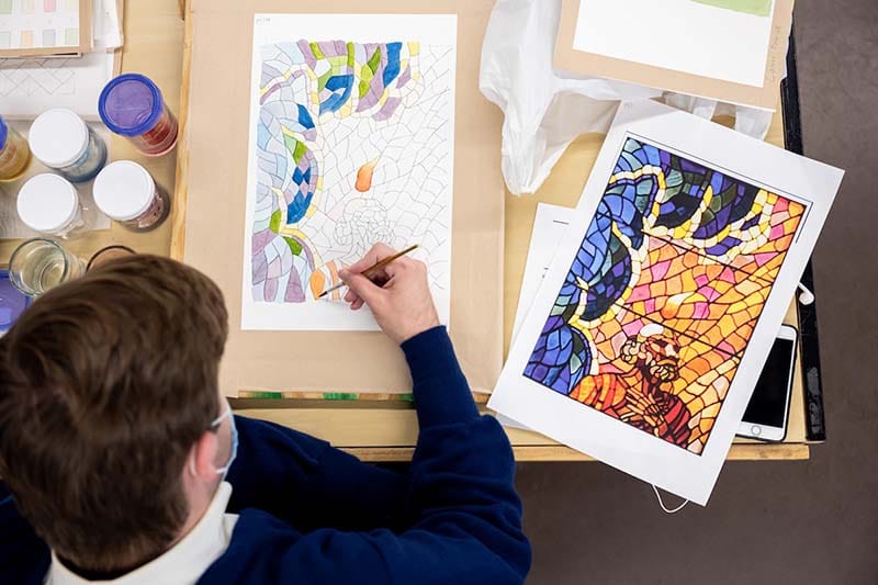 A student works on a watercolor of stained glass window depicting a saint.