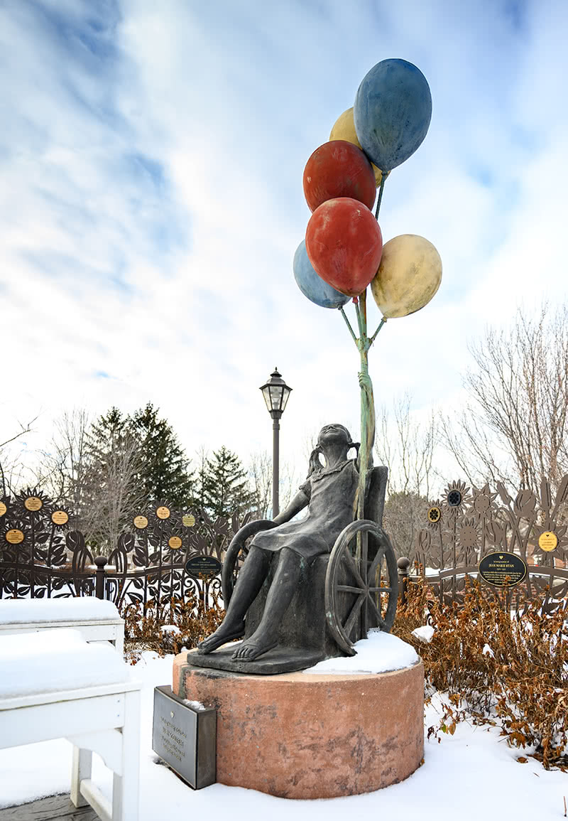 Statue of a child in wheelchair holding 6 colorful baloons. The statue is in front of Misericordia grounds.