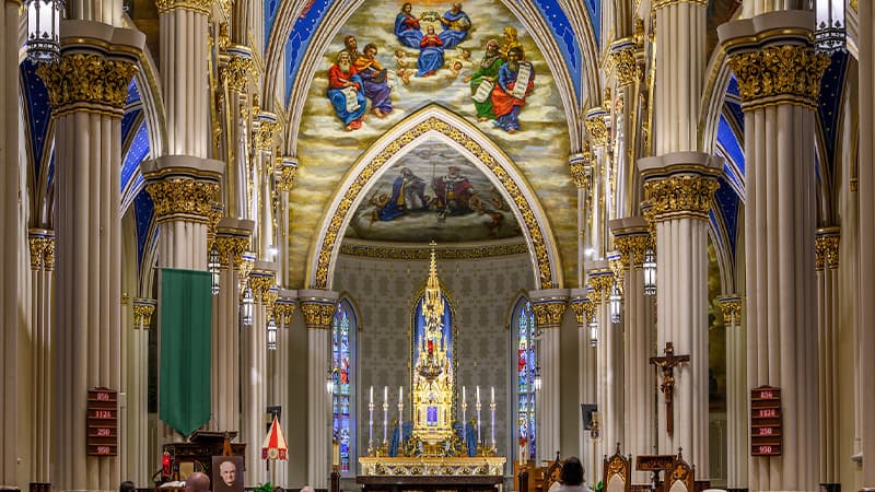 The Basilica of the Sacred Heart has ornate white columns with gold and blue details on either side of the nave. The tall, arched ceilings are covered in elaborate murals depicting heavenly scenes. A Catholic alter with three candles on each side of a tall golden tabernacle tower is the central focus of the image.