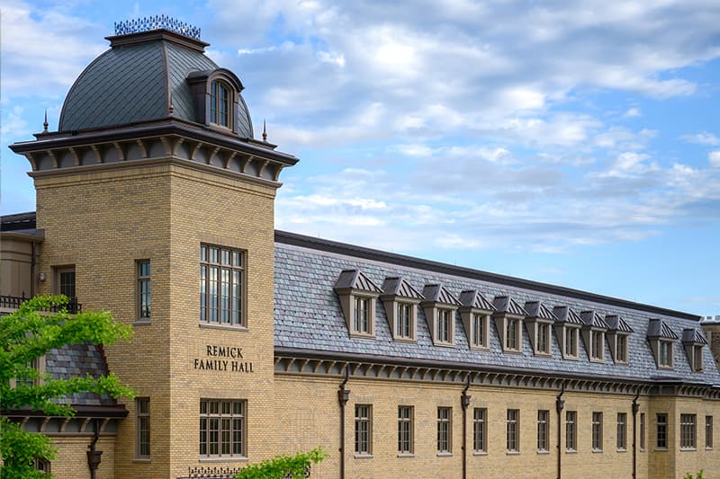 The upper floors of Remick Family Hall contrast a blue sky and white clouds. Yellow bricks of the wall are lined with a row of rectangle windows. The greyish-green clay tiles line the roof where another row of windows can be seen.