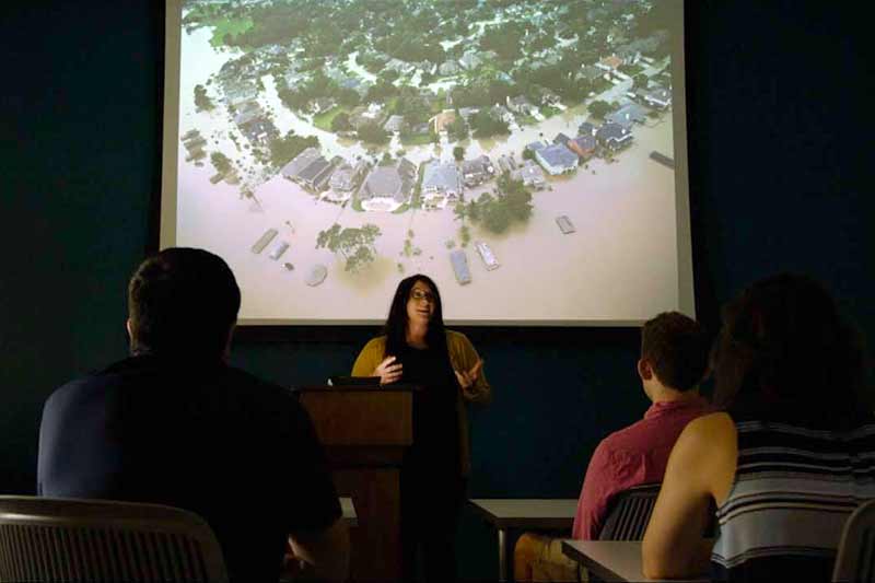 A woman wearing glasses speaks in front of a projector. Displayed on the projector is an area flooded by a hurricane.