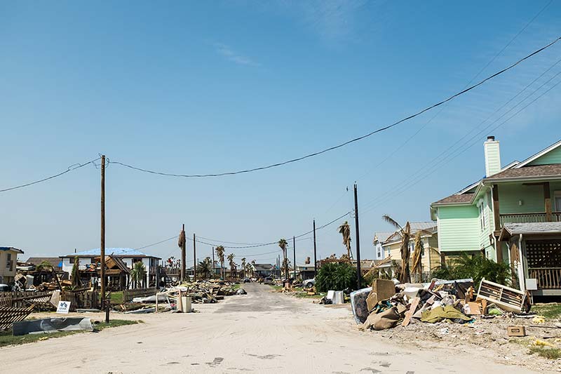 A look down the center of a road, piles of debris can be seen and wind-torn homes on both sides of a road.