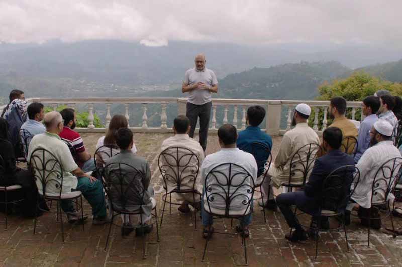 A group of people sit to listen to a professor talk in front of background of mountains.