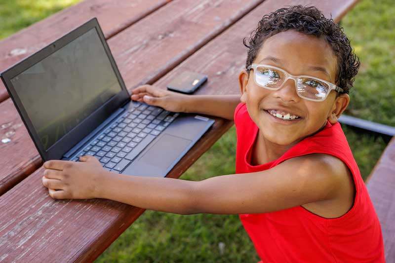 A young boy, wearing glasses, holds onto his laptop computer and smiles up at the camera.