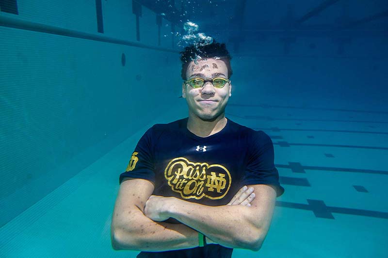 An underwater photo in a swimming pool of a male student wearing googles with his arms crossed wearing a shirt that reads Pass It On ND.