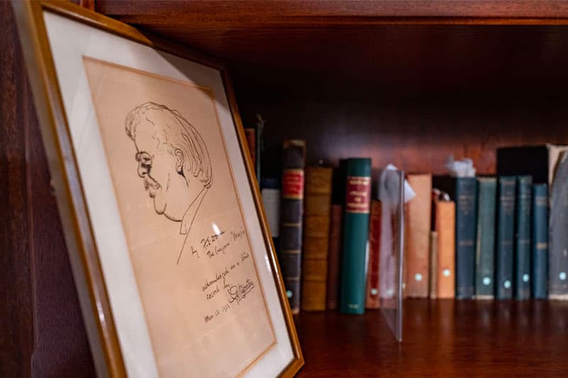 AA caricature of GK Chesterton in a frame, sitting to next to a shelf with all his books.