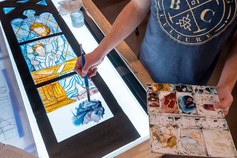 A students paints a luminaire — a mockup of the window painted onto clear acetate and framed, so that light can shine through it 